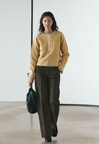 Mosterdkleurige geribde trui met een knoopsluiting bij de hals, gecombineerd met een hoge taille broek in bruin; houdt een zwart tas met structuur vast; eenvoudig ontwerp.