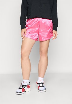 Personne portant un short de sport rose brillant, un sweat-shirt noir, des chaussettes blanches et des baskets blanches, rouges et noires, debout sur un sol gris.