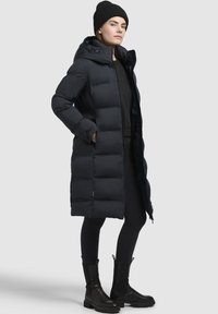 Dlhý čierny páperový kabát s kapucňou, s prešívaným dizajnom a dvoma bočnými vreckami. Skombinovaný s čiernymi legínami a členkovými topánkami.