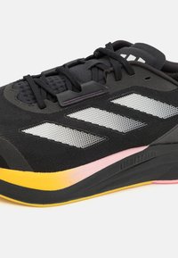 Zwarte sportieve schoen van meshmateriaal, met grijze accenten en een gele buitenzool. Bevat drie witte diagonale strepen en een roze tussenzool.