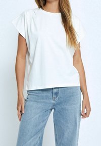 Bílá bavlněná tričko s krátkými rukávy, uvolněný střih a jemnými sklady na ramenou. Komplementováno světle modrými džínami.