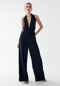 Tamsiai mėlynas marinis kombinezonas su giliais V formos iškirpte, plėstomis kojomis ir tekstūriniu paviršiumi. Turi auksinį akcentą juosmens srityje ir minimalias dizaino detales.