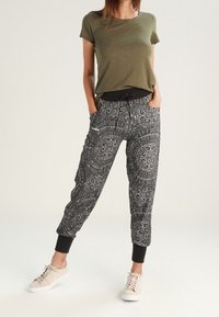 Zielona oliwkowa koszulka zestawiona z czarnymi spodnimi dresowymi z wzorem, które mają regulowany pas, zwężane mankiety i wzór w spirale. Całość uzupełniają białe sneakersy.