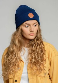 Granatowa czapka z dzianiny z okrągłą pomarańczową skórzaną łatką z przodu; model ma na sobie żółtą kurtkę na białą koszulę. Jasne, kręcone włosy.