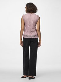 Kvinne sett bakfra iført en ermeløs rosa blonde-topp, sorte bukser med vide ben og svarte sandaler med hæl på hvit bakgrunn.