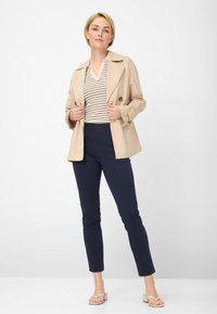 Beige Doppelkindertrenchcoat über einem gestreiften Oberteil, kombiniert mit schmal geschnittenen navy Hosen und beigen Flip-Flop Sandalen. Glatte Textur und maßgeschneiderte Passform.