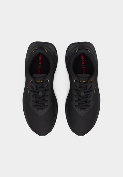 Paire de baskets noires Hugo avec œillets dorés, lacets noirs et logo rouge Hugo à l'intérieur des chaussures, vues de dessus sur un fond gris.