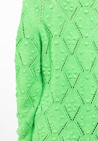 Maglione verde lavorato a maglia con un design testurizzato caratterizzato da motivi a diamante e palline in rilievo. Materiale leggero con dettagli di cuciture aperte.