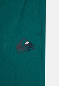 Tealfarget genser med myk tekstur, med et mørkt logo design av en bølge og et fjell. Inkluderer en kengurulomme.