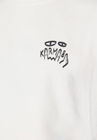 Weißer Stoff mit schwarzer Grafik, die zwei Augen über dem Wort "KARMA" zeigt, integriert mit handgezeichneten Fingern.