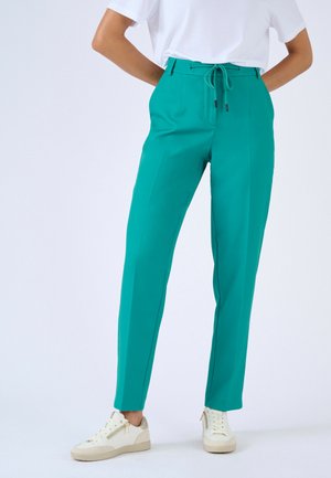 Personne portant un pantalon fuselé bleu sarcelle avec passants de ceinture et des baskets blanches, debout les mains derrière le dos devant un fond uni.