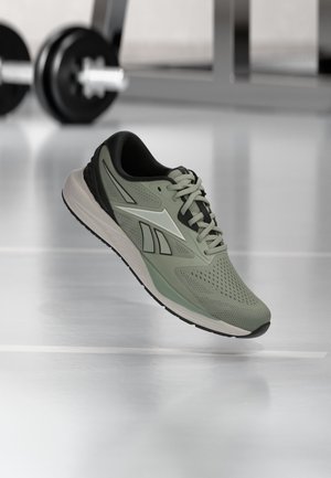 Chaussure de sport verte et noire avec un motif blanc, lacets noués, présentée sur un fond flou de salle de sport avec des haltères.