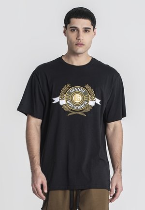 Mladý muž nosiaci čierne oversized tričko so zlatým a bielym logom Gianni Kavanagh uprostred na hrudi.