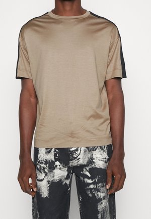 Mann trägt beige kurzärmliges Hemd mit schwarzen Schulterstreifen und schwarz-weiß abstrakt gemusterte Hose, steht vor einfarbigem Hintergrund.