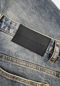 Denim jeans met een lichtblauwe wassing, voorzien van een zwart leren label met de tekst "BUCK BENNIS" en contrasterende gele stikdetails.
