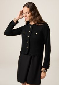 Zwarte cropped cardigan met gouden knopen, twee voorkanten zakken en een gestructureerde rand. Gemaakt over een zwarte jurk, met een gladde stofafwerking.