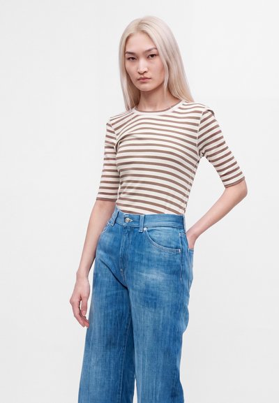 Jeune femme aux longs cheveux blonds portant une chemise rayée beige et blanche et un jean bleu taille haute à jambes larges, debout avec une main dans la poche.