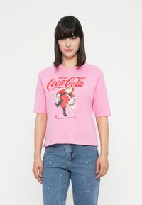 Žena oblečená v ružovom tričku Coca-Cola so Santa Clausom, ktorý drží fľašu a malým jeleňom, v kombinácii s modrými džínsami s jemnými kvetinovými vzormi.