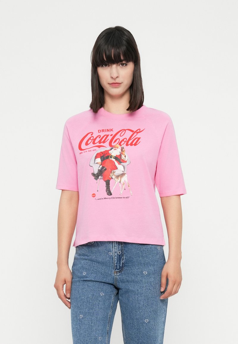 Žena oblečená v ružovom tričku Coca-Cola so Santa Clausom, ktorý drží fľašu a malým jeleňom, v kombinácii s modrými džínsami s jemnými kvetinovými vzormi.