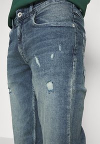 Jeans de mezclilla azul deslavado con parches desgastados y bordes deshilachados. Presenta un diseño clásico de bolsillos y presillas para el cinturón. Superficie texturizada con costuras visibles.