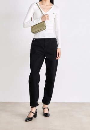Person trägt ein weißes Langarm-Top, schwarze Hose, schwarze Schuhe mit Absatz und hält eine olivgrüne Schultertasche.