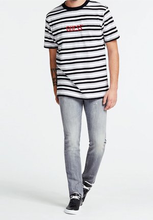 Mann trägt ein schwarz-weiß gestreiftes Guess-T-Shirt, hellgraue ausgewaschene Jeans und schwarze Sneaker mit weißen Schnürsenkeln, steht vor weißem Hintergrund.