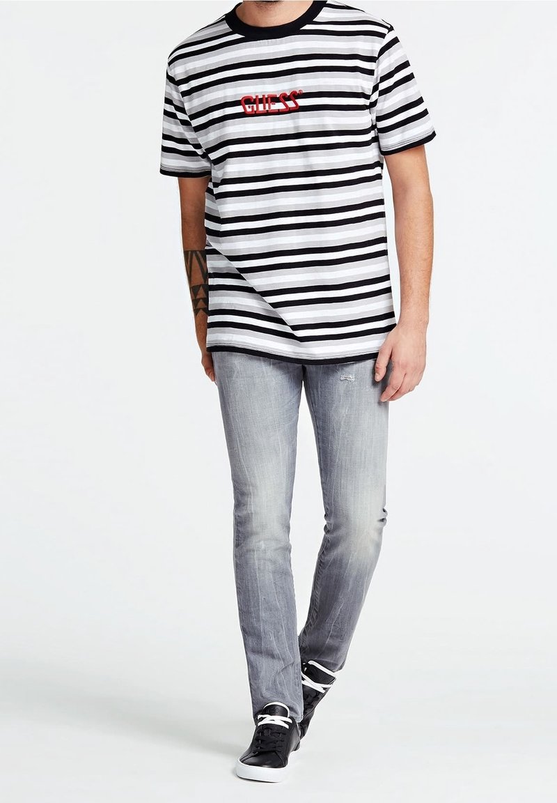 Homme portant un t-shirt Guess rayé noir et blanc, un jean gris clair délavé et des baskets noires à lacets blancs, debout devant un fond blanc.