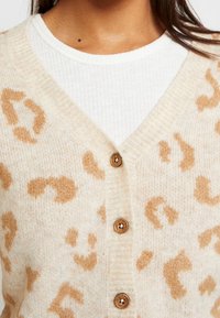 Cárdigan beige con estampado de leopardo en marrón claro. Presenta un escote en V, botones de madera y una tela suave y texturizada.