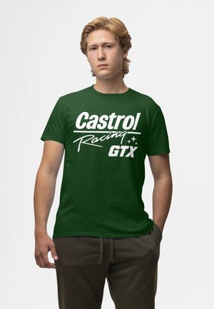 Ung mand med blondt hår iført en mørkegrøn "Castrol Racing GTX" t-shirt og mørke bukser, stående med den ene hånd i lommen.