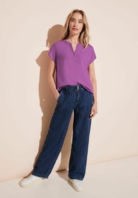 Blusa viola a maniche corte con scollatura a V, abbinata a jeans scuri a gamba larga e sneakers bianche. Design semplice, texture del tessuto liscia.