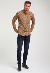 Rjav srajc s geometrijskim vzorcem, v kombinaciji s temno modrimi slim-fit kavbojkami in mornarskimi supergami. Srajca ima ovratnik in dolge rokave.