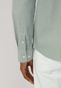 Camicia a quadri verde e bianca con maniche lunghe. Presenta bottoni chiari e un orlo segmentato, abbinata a pantaloni bianchi.