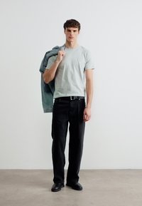 Νεαρός άνδρας που στέκεται κρατώντας ένα τζιν μπουφάν στον ώμο, φορώντας ένα ανοιχτό γκρι μπλουζάκι, μαύρα τζιν και μαύρα παπούτσια.