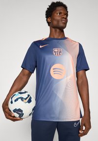 Nike football t-shirt in navy with orange accents, featuring a Barcelona logo, Spotify circle, and wave patterns. Model holding a football.