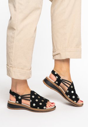 Pies con sandalias negras con lunares plateados, tiras elásticas y pantalones de pana color marrón claro con puños remangados sobre un fondo blanco.