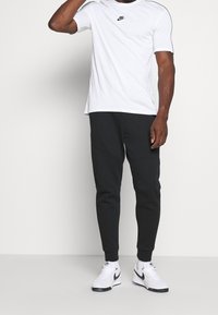 Bílá tričko Nike s černým logem, zkombinované s černými tepláky s zakřiveným designem a bílými teniskami s černými akcenty.