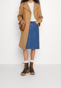 Abrigo de camello con grandes solapas, suéter de punto color crema, falda de mezclilla y botas marrones con cordones, suela gruesa y acentos de hardware dorado.