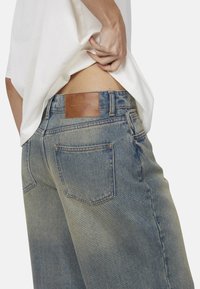 Jeans in denim blu con una finitura sbiadita, caratterizzati da una toppa posteriore in pelle marrone, cinque tasche e dettagli di cucitura in evidenza.