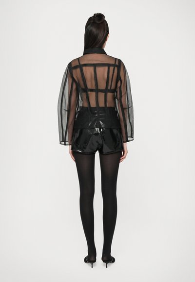 Čierna priesvitná bunda so štruktúrovanými detailmi cez lesklé čierne vysoko pásové šortky, spárované s čiernymi pančuchami a lodičkami na vysokom podpätku.
