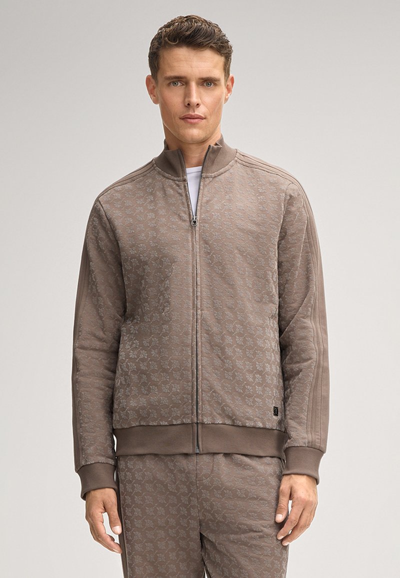 Braune Zip-Up-Trackjacke mit dezentem Blumenmuster, gerippten Bündchen und Saum, weichem Material und taillierter Silhouette. Passende Hose abgebildet.