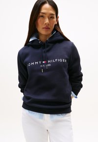 Granatowa bluza z kapturem z białym i czerwonym logo, z regulowanym sznurkiem w kapturze, miękkiego materiału, warstwowa z jasnoniebieską koszulą z kołnierzykiem pod spodem.