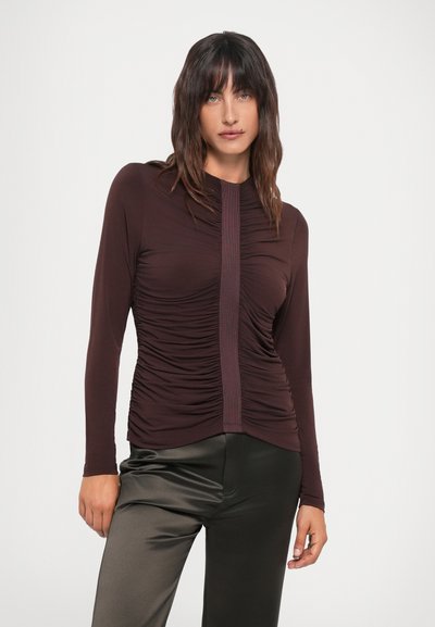 Long-sleeve, ruched top in dark burgundy with a central panel. The fabric is smooth, complemented by fitted, shiny black pants.
