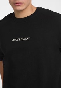 Sort bomulds T-shirt med rund halsudskæring og broderet "GUESS JEANS" logo i hvid. Korte ærmer og klassisk pasform.