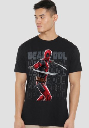 Young man wearing black t-shirt featuring Deadpool in red suit holding two swords, with repeated "Deadpool" text in background.