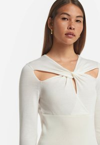 Vit långärmad topp med knutdetalj och urtagna axlar, med en slät textur och en figurnära silhuett.