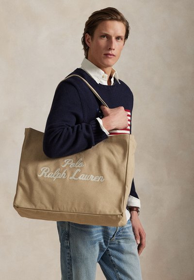 Άνδρας που φοράει σκούρο μπλε πουλόβερ με αμερικανική σημαία, κρατώντας μεγάλη μπεζ τσάντα ώμου Polo Ralph Lauren μπροστά σε απλό φόντο.