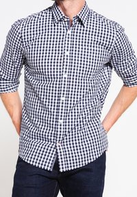 Uomo che indossa una camicia a quadri blu e bianca con le maniche arrotolate e jeans blu scuro, con le mani posate dietro la schiena.