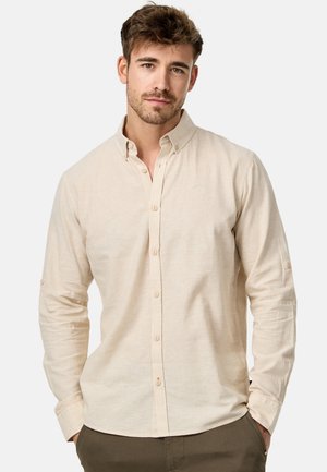 Camisa abotonada de color beige claro con una textura sutil, de manga larga y detalle de botón para enrollar. Presenta un pequeño acento bordado sobre el bolsillo.