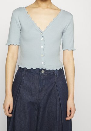 Kobieta ubrana w jasnoniebieską koszulę z krótkim rękawem zapinaną na guziki z falistymi brzegami oraz w ciemnoniebieskie, szerokie spodnie z wysokim stanem.