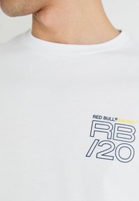 Balts kokvilnas T-krekls ar tumši zilu un dzeltenu grafiku uz krūtīm, kurā rakstīts "RED BULL® RACING RB/20" modernā, uzsvērta fontā.